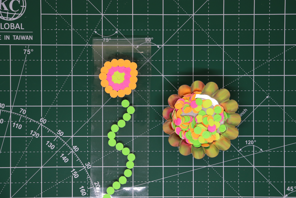 A container of colorful paper dots, by packing tape with a green paper dot stem and yellow, pink, and orange paper dots in concentric circles making the beginning of a flower