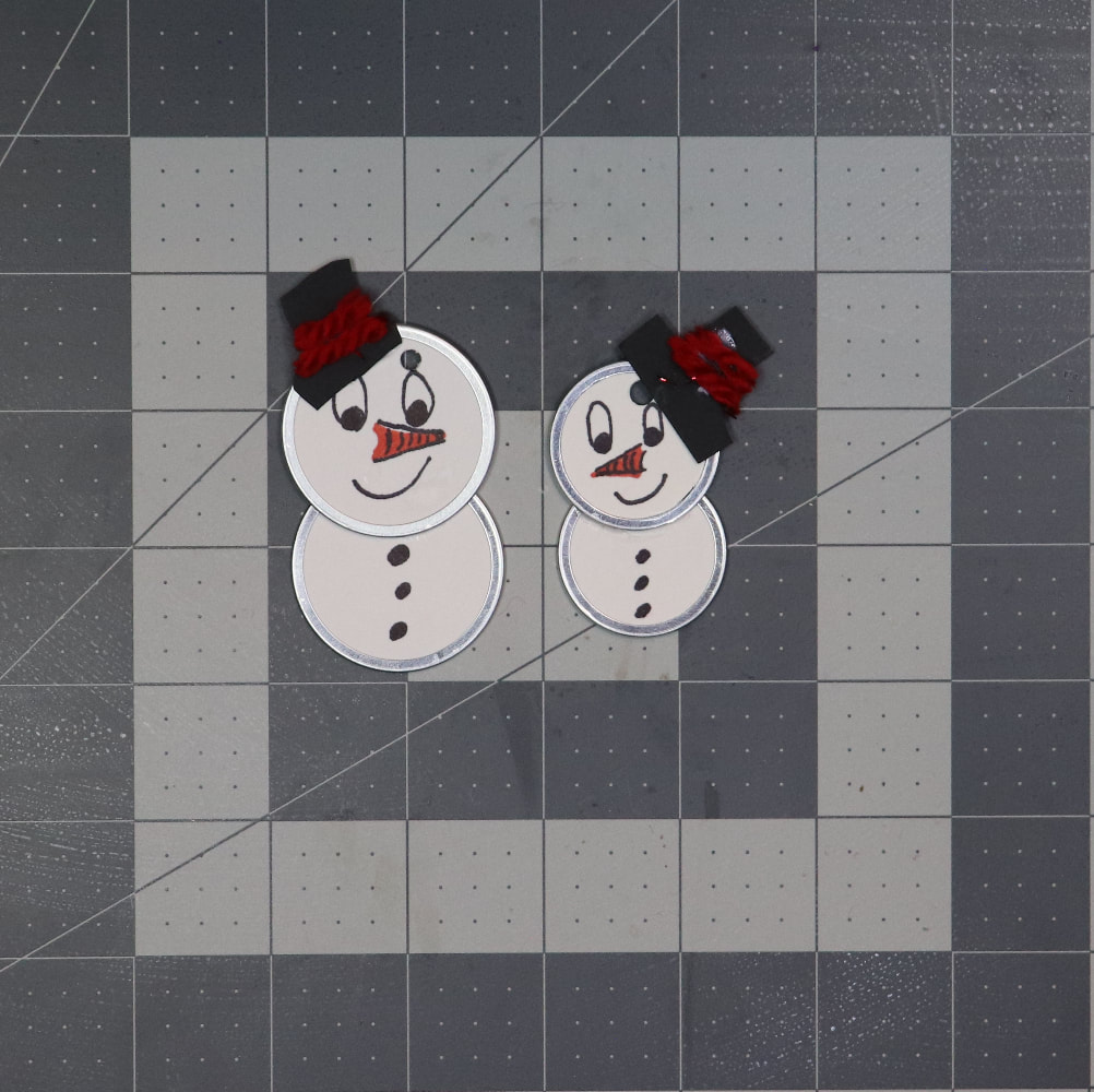 Two snow people ornaments made out of metal rim key tags. They have black hats tilted to one side with red yarn glued down where a hat band would be.