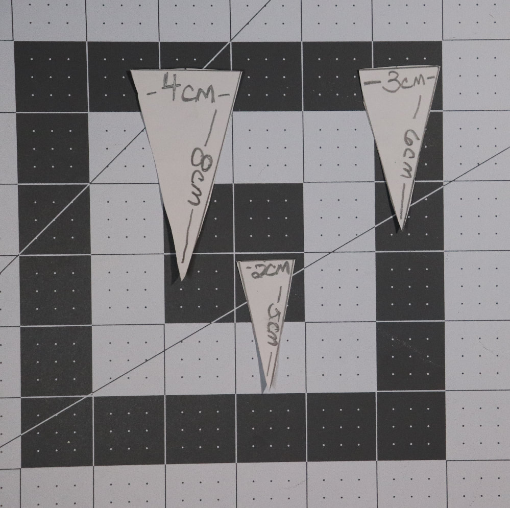 A cutting mat with three triangles cut out of index card on it. The measurements are written on the triangles, one across the top and one down one side: 4cm and 8cm on the largest, 3cm and 6cm on the medium, and 2cm and 5 cm on the smallest.d