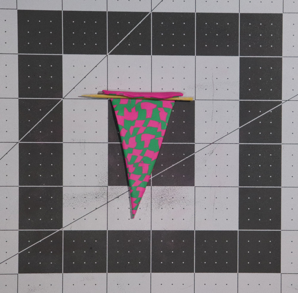 A checkerboard patterned triangle of clay in green and pink with a toothpick at the top of the triangle (the shortest side) the clay is slightly curved over the toothpick. 