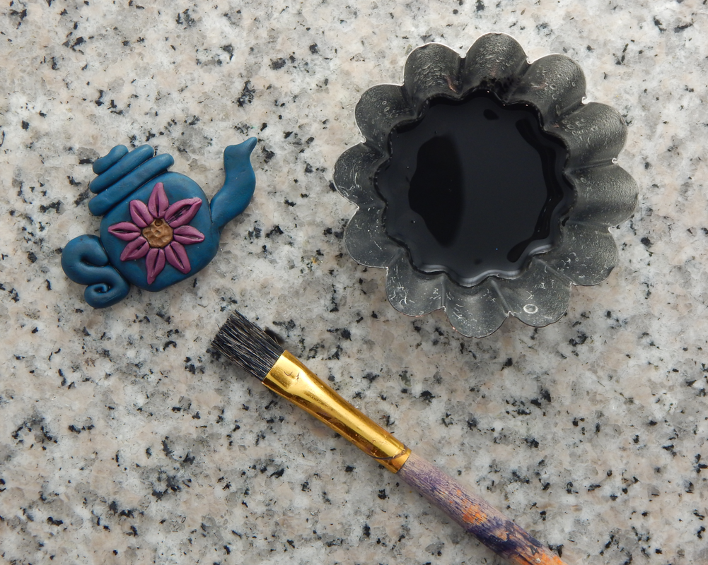 A blue teapot bead with pink flower is in one corner. In the other corner a fluted metal tin holds diluted acrylic paint. At the bottom there is a stiff bristled paintbrush.