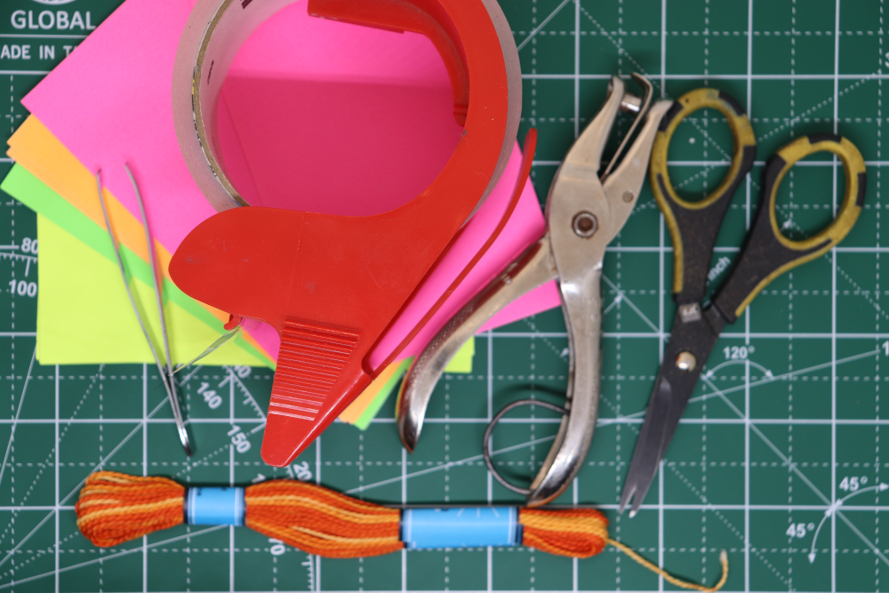 A picture of supplies: a fan of colored index cards, packing tape, a hole punch, scissors, tweezers, and embroidery thread