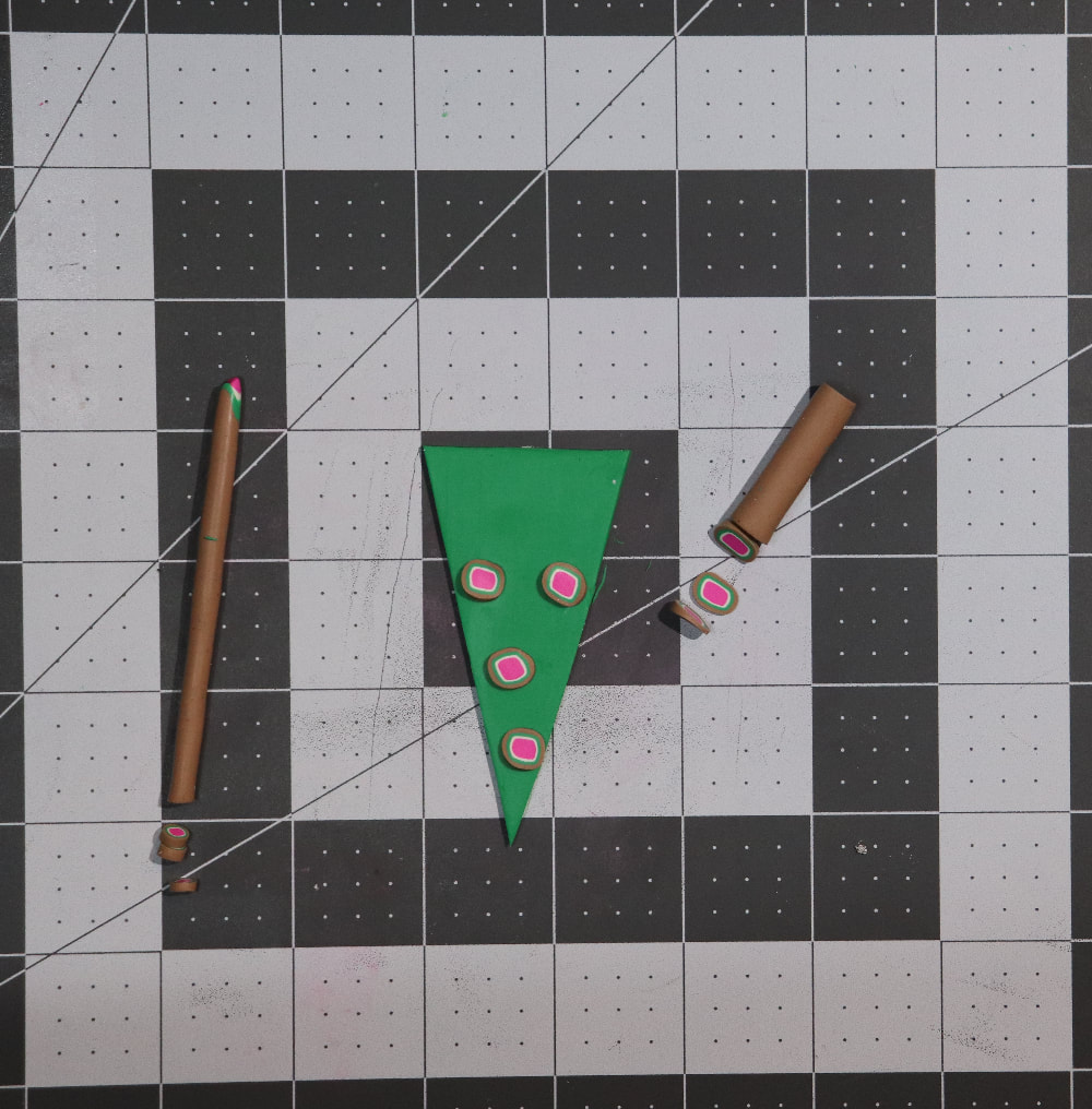 A green triangle of clay with pink, white, green, and brown polka dot canes on it. The canes are on the side showing several slices cut.