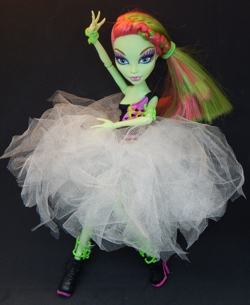 A green skinned fashion doll with pink and one curving in front of her torso. She wears a pink, green, and black one shouldered dress and a fluffy white tutu with chunky black boots with pink soles.