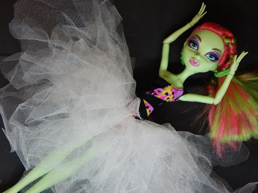 A green skinned fashion doll with pink and green hair lies on a black background with her arms reaching towards her head. She is wearing a black and pink one armed dress and a white tutu at her waist. 