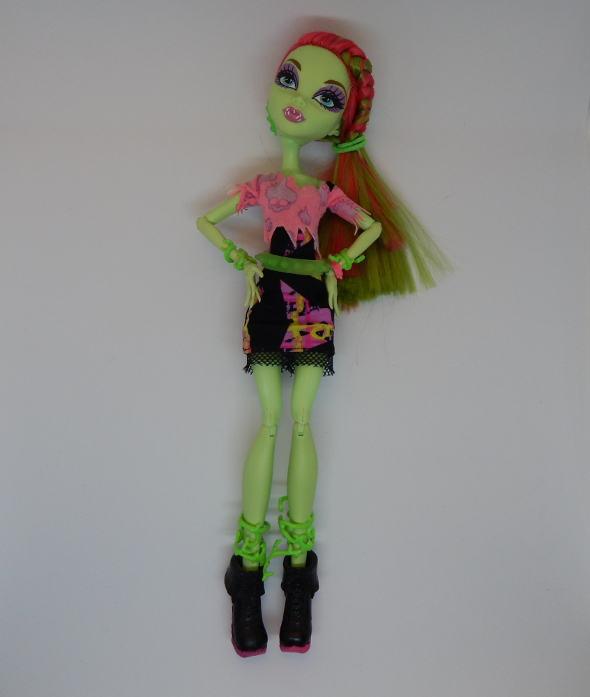 A green skinned fashion doll with pink and green hair on a white background. Her hands are on her hips and she is wearing chunky black boots. 