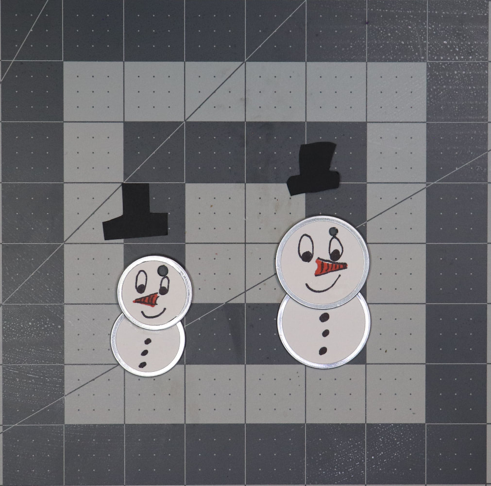 Two metal rim tags with faces and buttons drawn on so they look like snow people. There are hat shapes cut out of black paper on the cutting mat above them. 