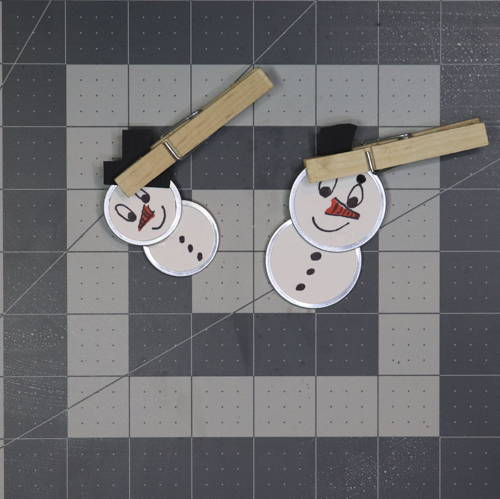 Two metal rim tag snow people ornaments. They have a black paper hat connected to the top with a clothes pin securing it so the glue can dry. 