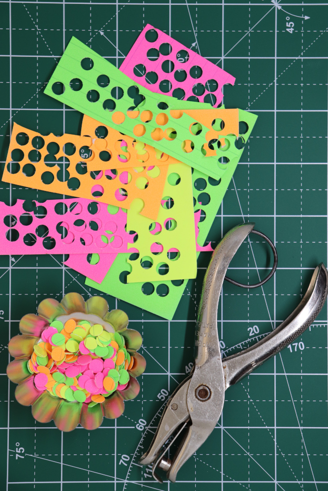 A few different pieces of paper in different colors with many punch holes in them, a small silver tin with many different colored paper dots, and a silver hole punch