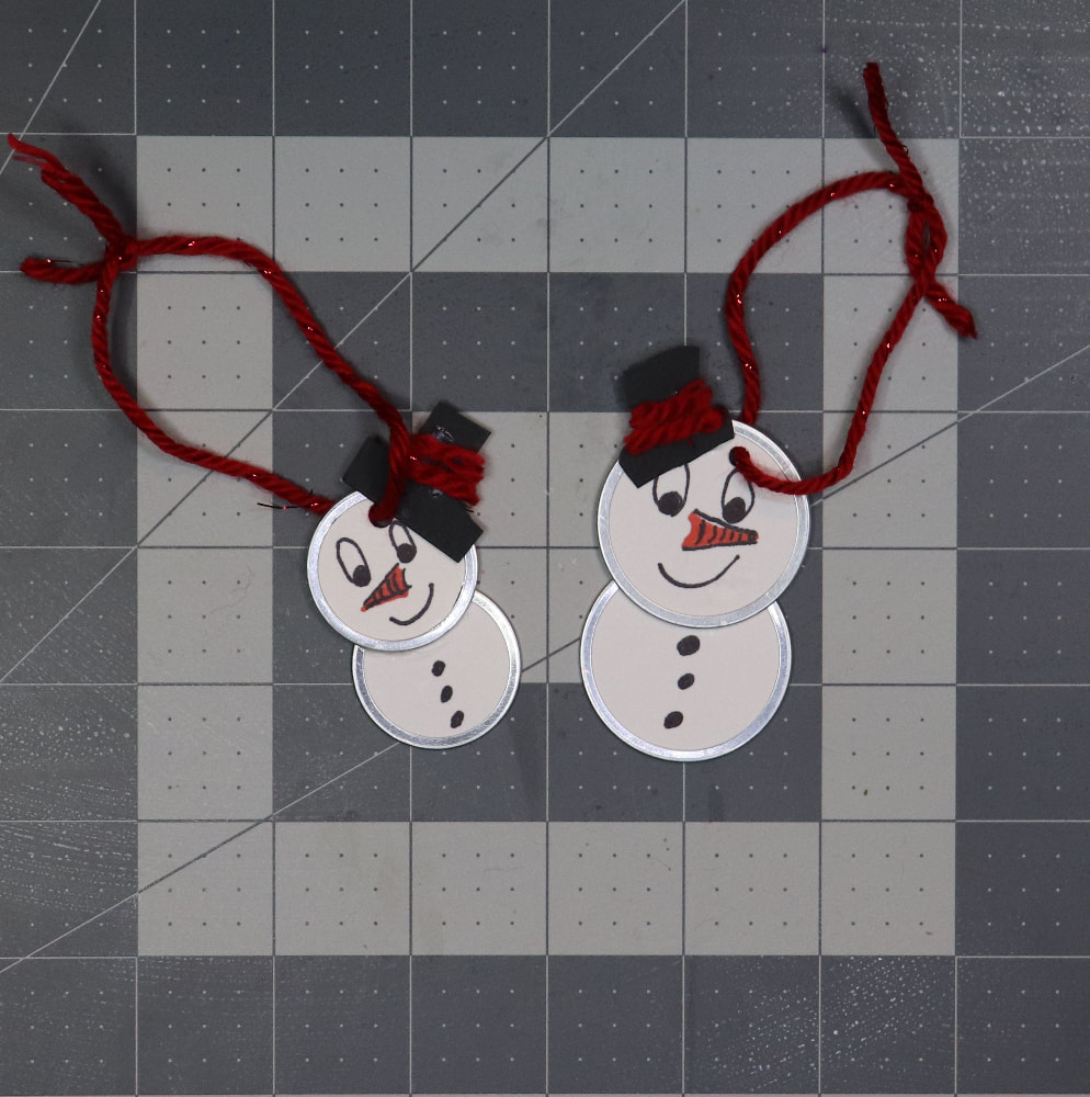 Two Snow people ornaments on a gray cutting mat. They have a loop of red yarn through the hole at the top and tied in a loop.
