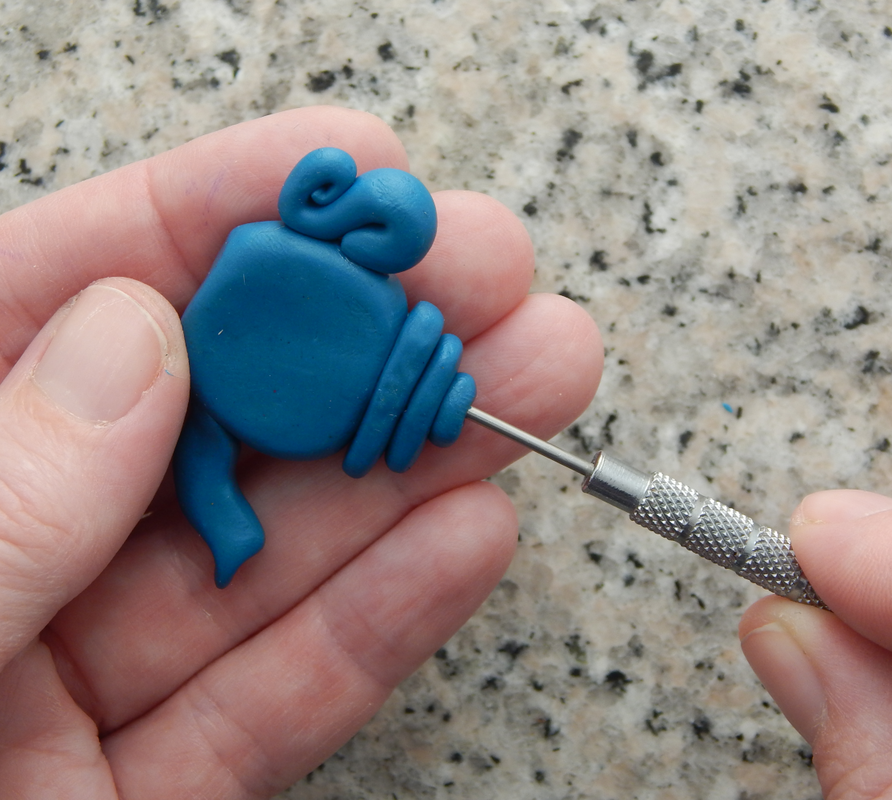 One hand holds a blue teapot bead, the other inserts a needle tool through the lid