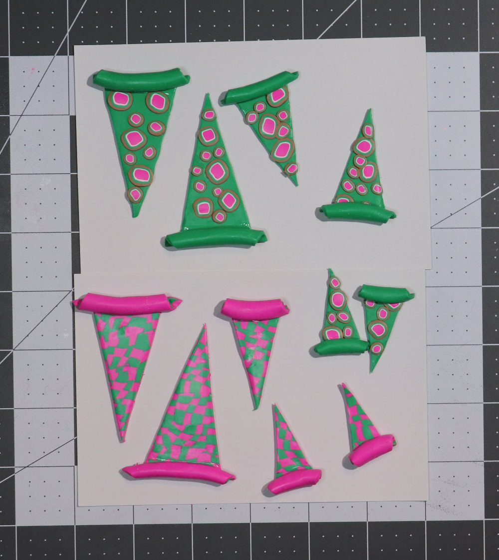 Patterned polymer clay triangles with a slightly curved tube at top on an index card. The triangles are green with pink, white, green, and brown polka-dots, and green and pink checkerboards. The triangles are of various sizes with one short side and two long equal sides. 
