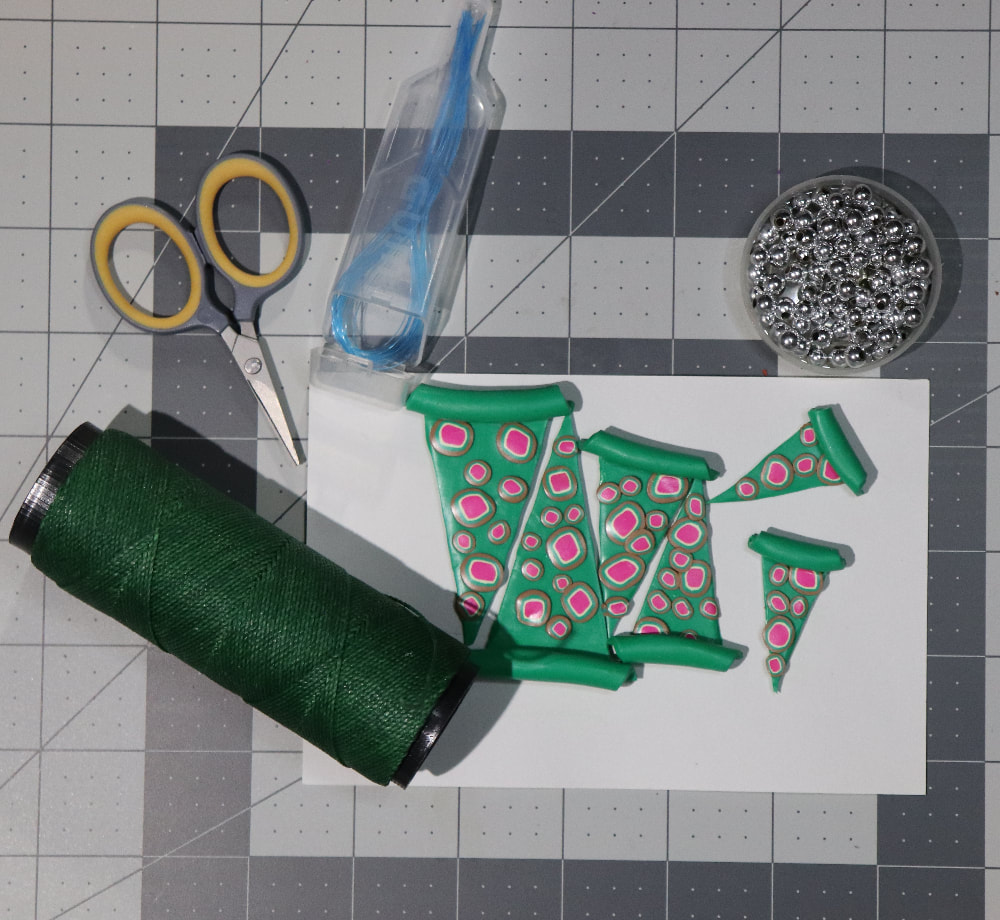 A gray cutting mat with stringing materials laid out on it: the baked green triangles, green cord, small scissors, floss threaders, and silver colored beads.