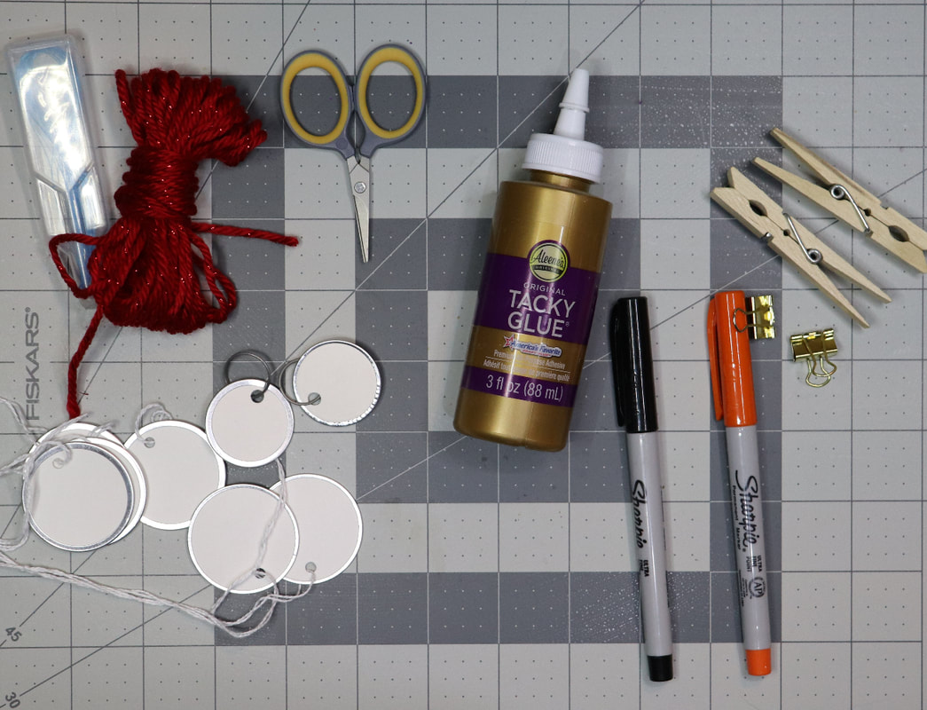 A light gray cutting mat with metal rim tags, scissors, markers, glue, clothes pin, binder clamps, and floss threaders laid out on it. 