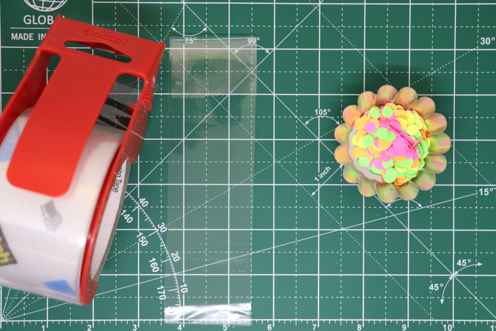 A roll of packing tape, a piece of the packing tape on the work surface, and a container of colorful paper dots
