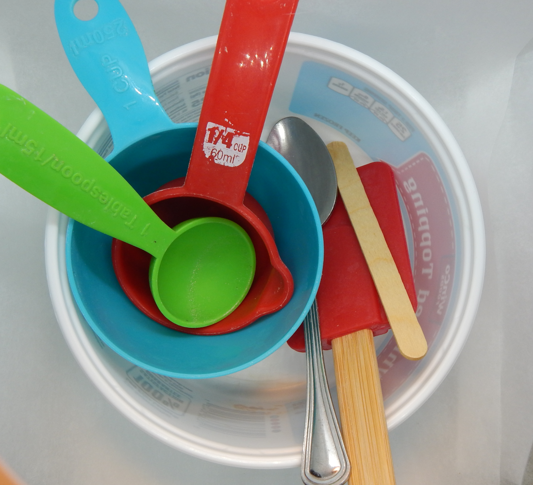 A white bowl filled with teal and red measuring cups, a green tablespoon, and a silver spoon, a popsickle stick, and a red spatula. 
